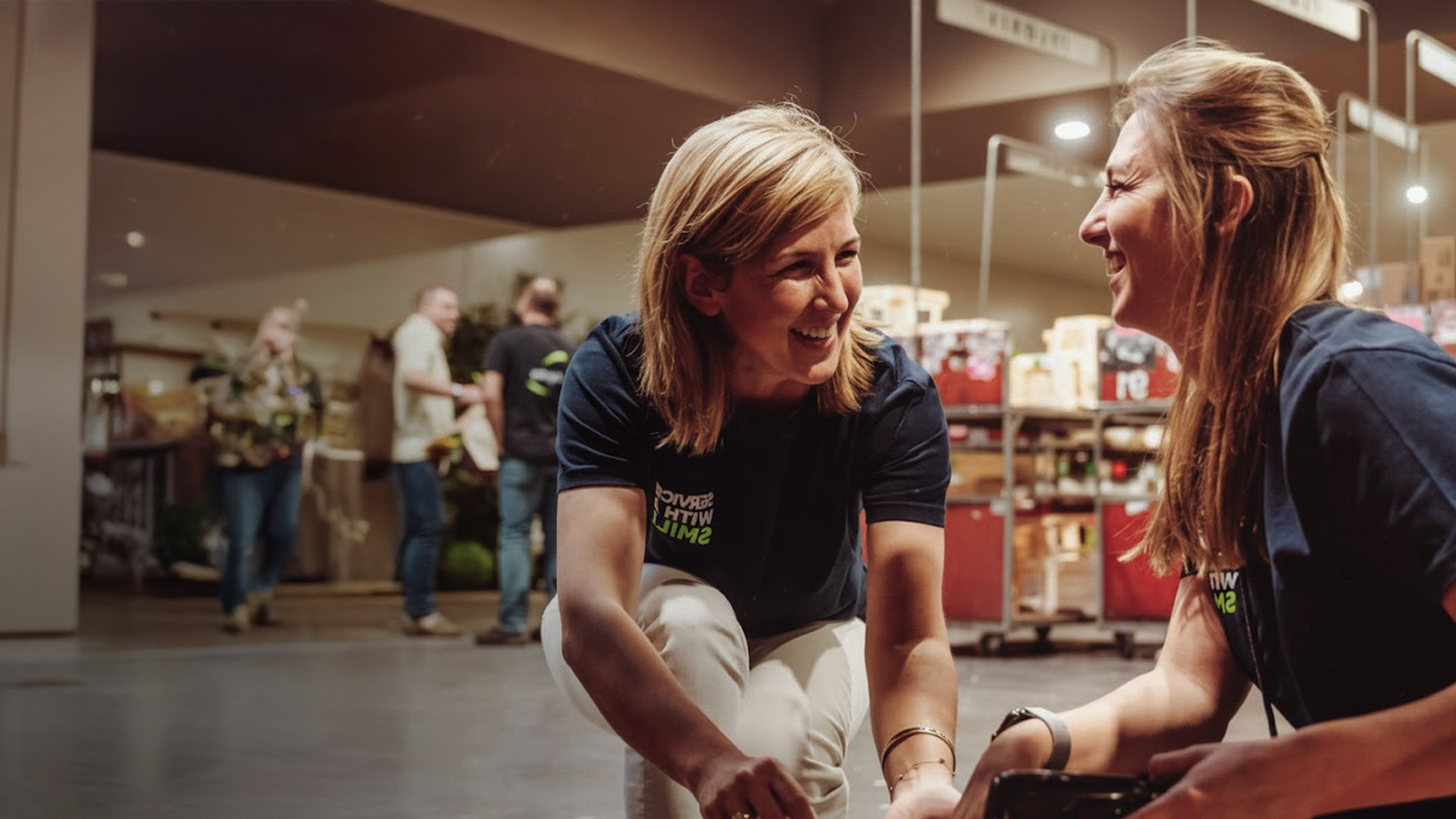 Twee medewerkers praten lachend in een magazijn, met karren en collega’s op de achtergrond.