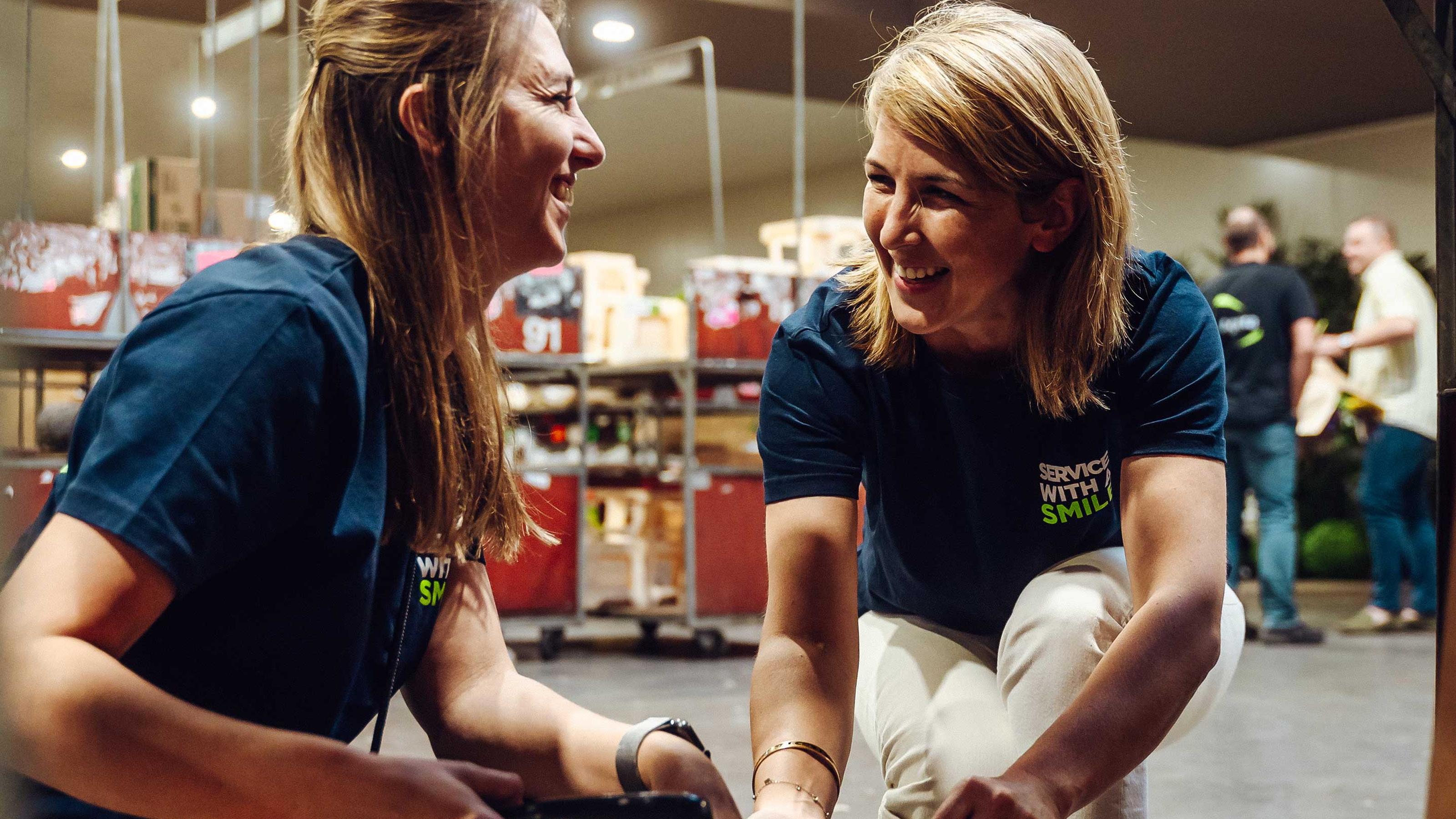 Twee vrouwen lachen terwijl ze bloemboeketten inpakken op een marktkraam.
