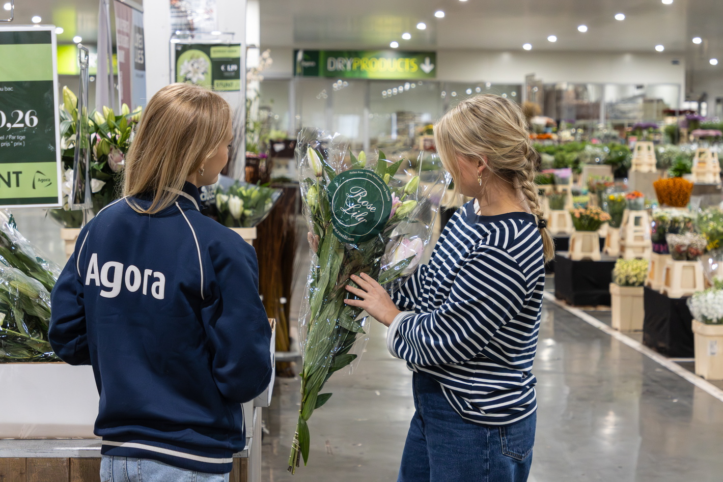 Twee vrouwen bekijken kamerplanten in een tuincentrum, bord “Agora” zichtbaar.