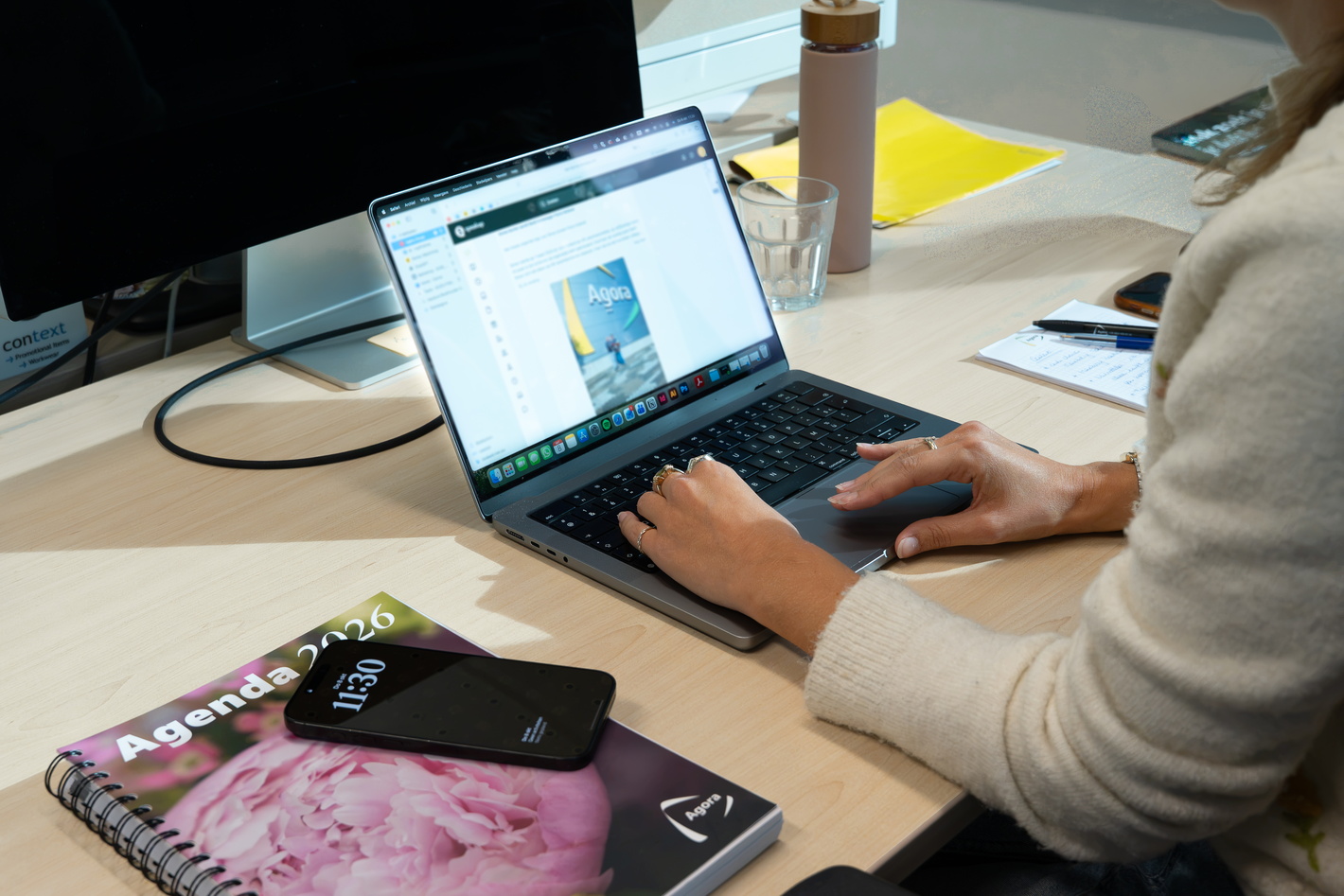 Persoon werkt op laptop aan bureau met notitieboek, smartphone en drinkbeker.