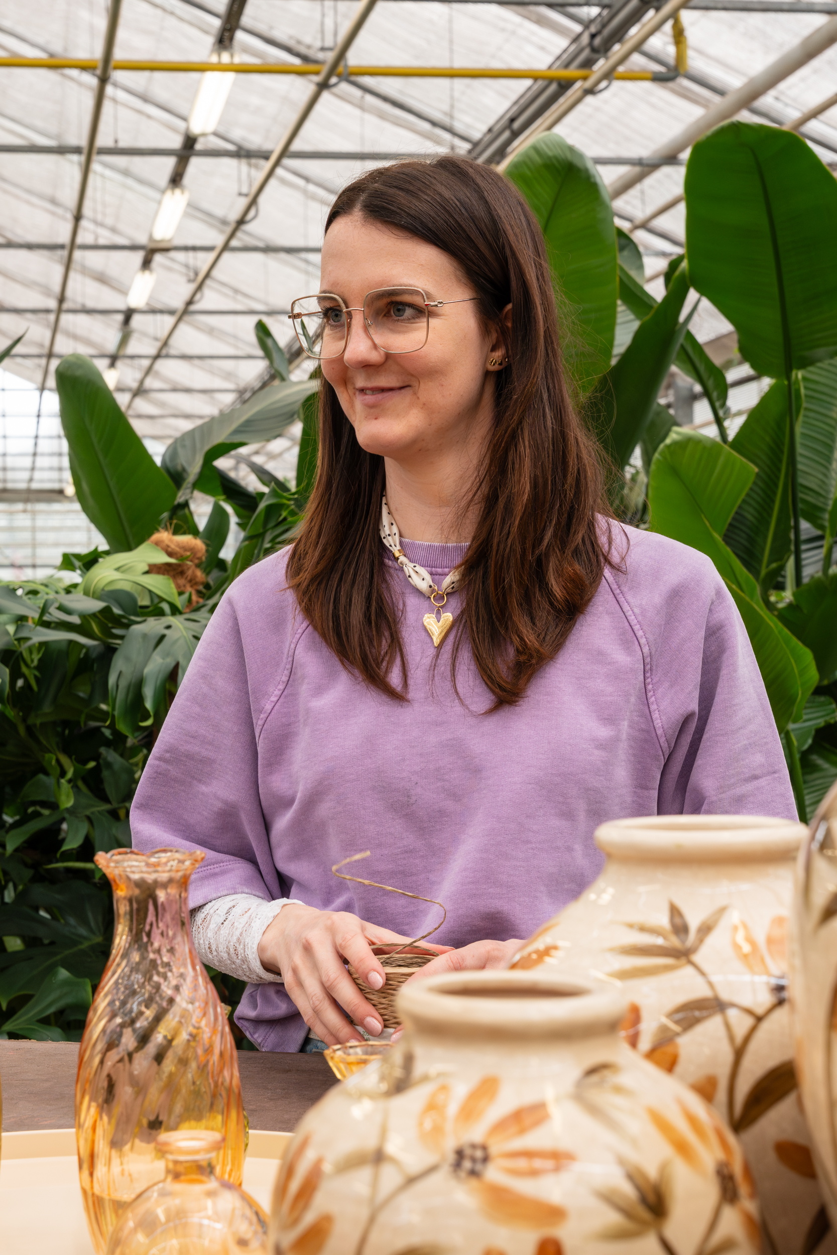 Vrouw in paarse trui zit in kas aan tafel met bloemenvaas en groene planten