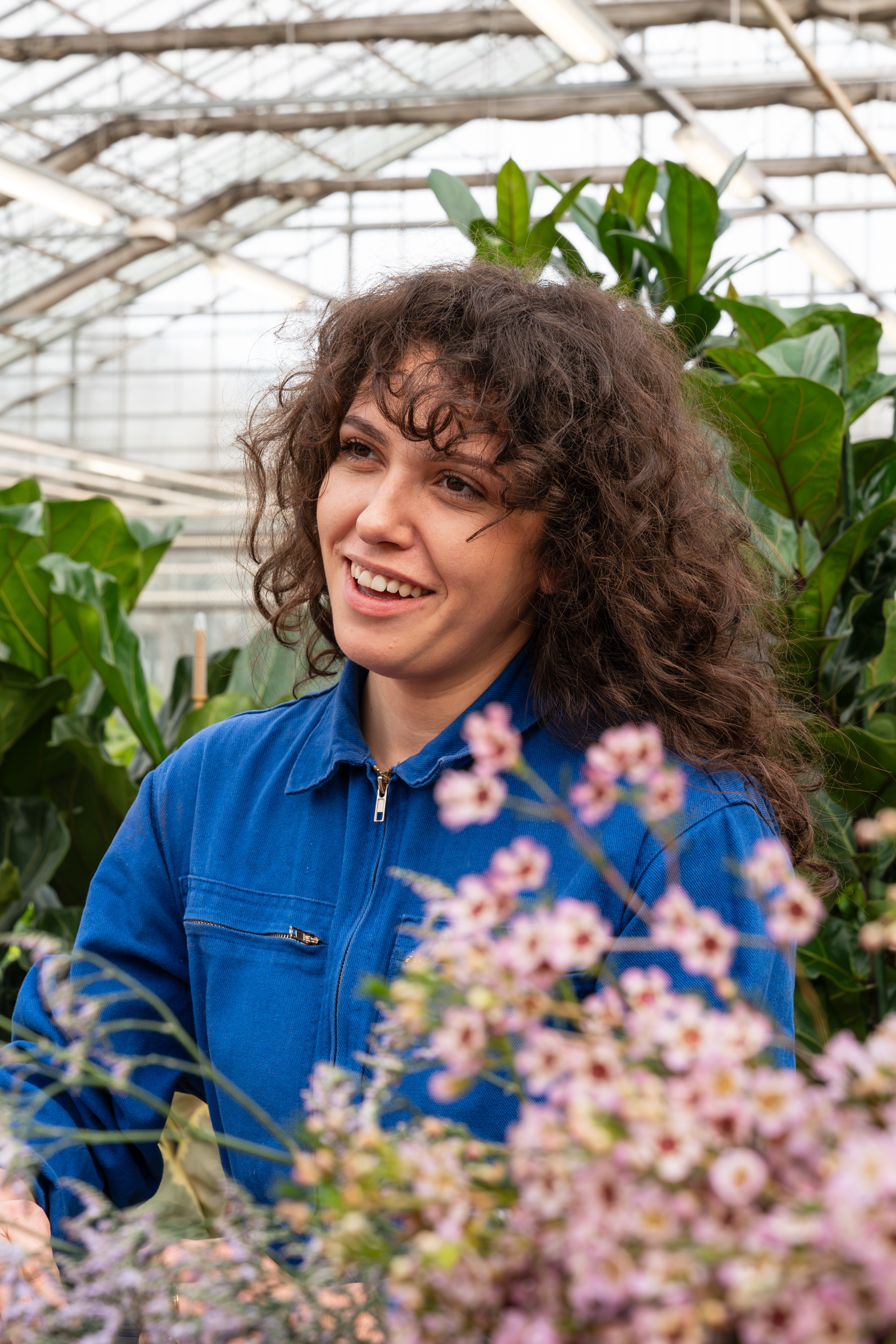 Vrouw met krullend haar in blauwe blouse tussen planten in een kas