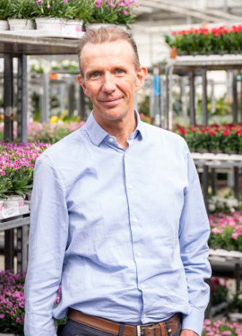 Man in lichtblauw overhemd in kas met rekken vol kleurrijke bloemen.