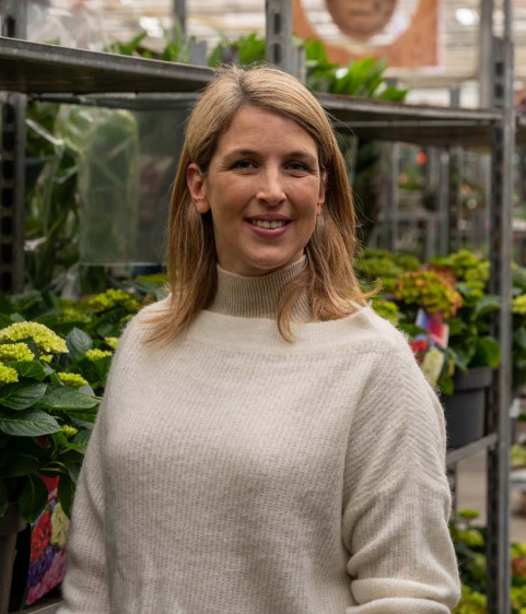 Vrouw in beige trui glimlacht tussen bloemen in een kas.