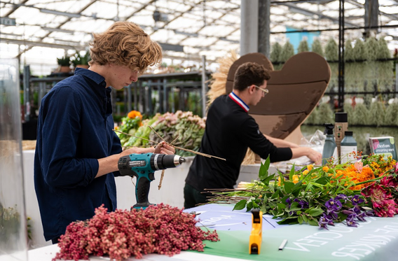 Twee mensen maken bloemstukken aan een tafel in een kas.