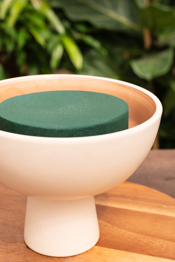 Witte keramische kom met groene inhoud op houten tafel, planten op de achtergrond.