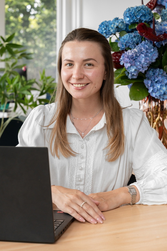 Vrouw glimlacht achter laptop aan houten tafel, met blauwe hortensia’s op achtergrond.