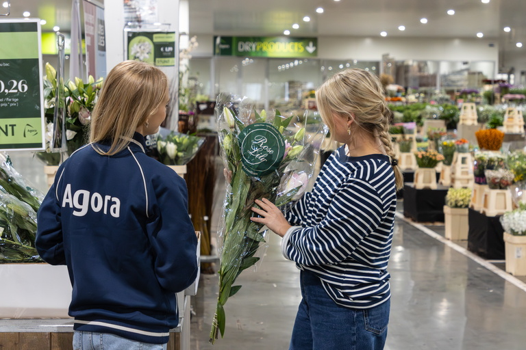 Twee vrouwen bekijken kamerplanten in een tuincentrum, bord “Agora” zichtbaar.