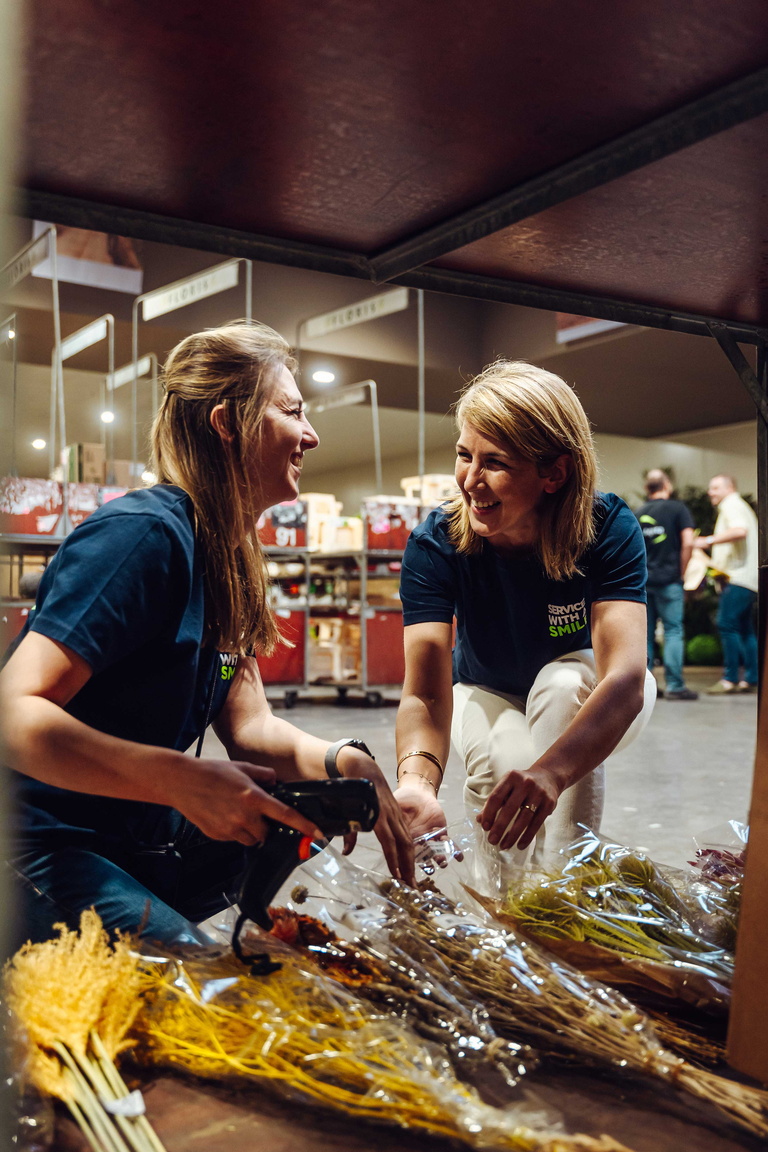 Twee vrouwen lachen terwijl ze bloemboeketten inpakken op een marktkraam.
