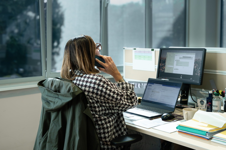 Vrouw op kantoor aan bureau met twee schermen, laptop en telefoon aan oor
