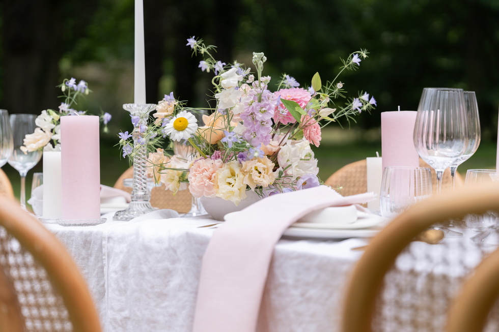 Gedekte buitentafel met bloemen, kaarsen en wijnglazen in zachte pastelkleuren.