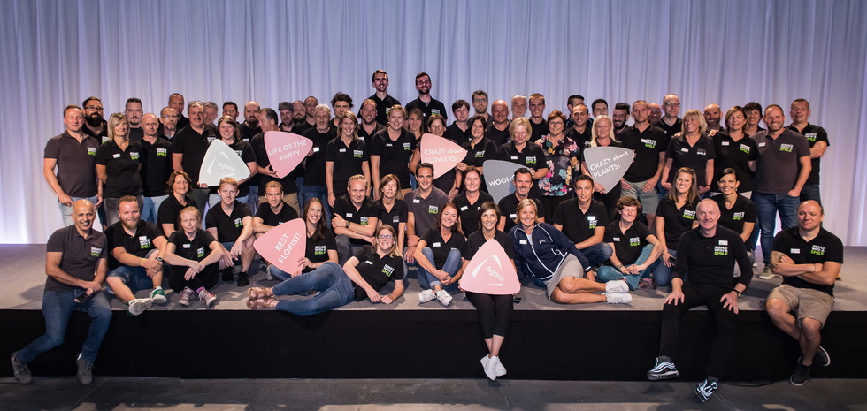 Grote groep mensen in zwarte kleding poseert op een podium met witte gordijnen.